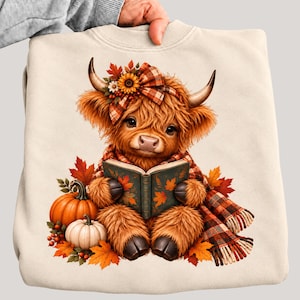 May include: A cream-colored sweatshirt featuring a graphic of a fluffy brown highland cow wearing a plaid scarf and a matching bow with a sunflower. The cow is holding an open book surrounded by orange and white pumpkins and autumn leaves.