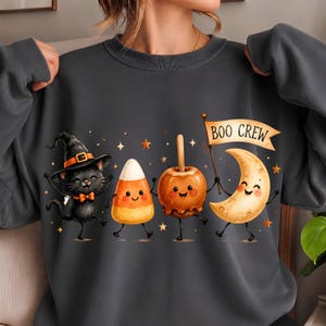 May include: A dark gray crewneck sweatshirt featuring a graphic of a black cat in a witch hat, a candy corn, a caramel apple, and a crescent moon with a face. The moon holds a flag with the text BOO CREW, surrounded by small orange stars.