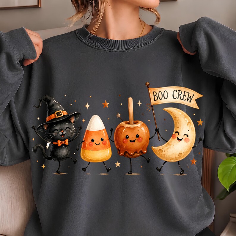 May include: A dark gray crewneck sweatshirt featuring a graphic of a black cat in a witch hat, a candy corn, a caramel apple, and a crescent moon with a face. The moon holds a flag with the text BOO CREW, surrounded by small orange stars.