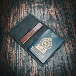 May include: Black leather passport holder with a brown leather insert and a blue passport. The passport has gold lettering and a crest. The holder is open, revealing the passport and insert on a dark wood surface.