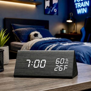 Op de afbeelding: Een driehoekige digitale wekker met een donkere houtnerfafwerking. Het display aan de voorzijde toont helderwitte LED-cijfers met de tijd 7:00, een luchtvochtigheid van 60% RH en een temperatuur van 26 F op een donkere achtergrond.