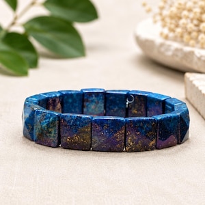May include: A stretch bracelet composed of rectangular, faceted stone beads in deep shades of blue, purple, and gold. The beads feature a mottled, galaxy-like appearance with metallic gold flecks distributed across the dark blue and violet surfaces.