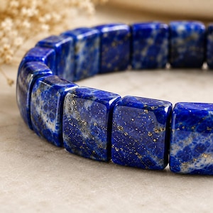 May include: A bracelet composed of rectangular, polished lapis lazuli beads. Each bead features a deep blue colour with visible natural inclusions of gold-coloured pyrite flecks and white mineral veining, arranged in a continuous circular formation.