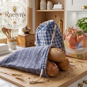 May include: Blue floral bread bag holding two loaves of bread, with a drawstring closure. Two additional drawstring bags are visible, one blue gingham and one patchwork. The Raelyn's Hive logo is in the upper left corner.