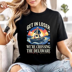 以下が含まれることがあります： 「GET IN LOSER」の文字と、ボートに乗った男性のイラストが描かれた黒いTシャツ。下には「WE'RE CROSSING THE DELAWARE」の文字。カラフルな夕焼けと水辺の風景が特徴的なグラフィックです。
