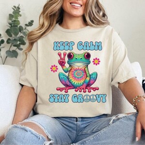 以下が含まれることがあります： カラフルなカエルのグラフィックがプリントされたベージュのTシャツ。カエルはタイダイ柄の腹をしており、ピースサインを出しています。「KEEP CALM STAY GROOVY」の文字が青い文字でカエルの上下に書かれています。