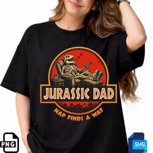 May include: Black t-shirt with a graphic featuring a skeleton dinosaur relaxing in a chair, holding a remote control. The design includes the text "JURASSIC DAD" and "NAP FINDS A WAY" in a stylized font, with a red and orange color scheme.