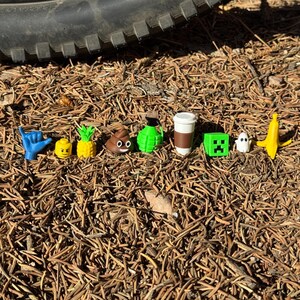 May include: A collection of small, colorful figurines, including a blue hand gesture, a yellow Lego head, a pineapple, a poop emoji, a green grenade, a coffee cup, a Minecraft block, a ghost, and a banana. The figurines are arranged on a bed of brown pine needles.