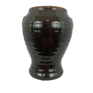 May include: A dark brown ceramic vase with a glossy finish. The vase has a wide, rounded body that tapers towards the base and a slightly flared rim. The surface reflects light, highlighting the curves and ridges of the design.