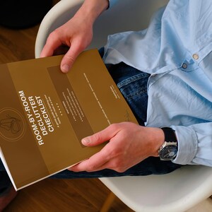 May include: A person holding a brown book titled "ROOM-BY-ROOM DECLUTTER CHECKLIST." The book has a circular logo and white text. The person is wearing a light blue button-down shirt and a watch with a black band.