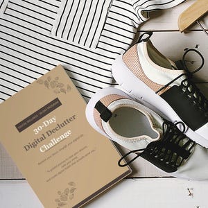 May include: A lifestyle flat lay featuring white and black trainers with pink detailing, a beige book titled "30-Day Digital Declutter Challenge", a striped top, and dark green trousers. The items are arranged on a white wooden surface.