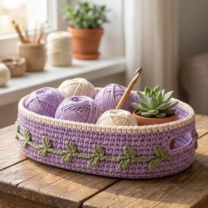 May include: An oval-shaped, light purple crocheted basket with integrated handles and a decorative green vine pattern stitched around the exterior. The basket contains several balls of lavender and cream yarn, a wooden crochet hook, and a small potted succulent.