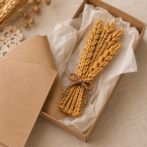 Può includere: Un mazzetto di spighe di grano lavorate all'uncinetto in giallo dorato, legato con un sottile fiocco di filo marrone, riposto all'interno di una scatola regalo rettangolare in cartone marrone foderata con carta velina bianca.