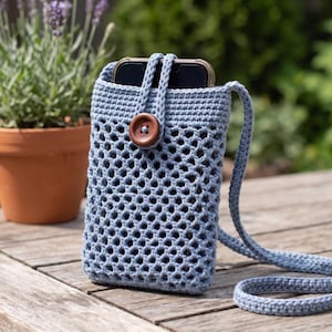Può includere: Una borsa a tracolla per telefono all'uncinetto blu chiaro con chiusura a bottone marrone. La borsa ha un design intrecciato e una tracolla abbinata. Uno smartphone è visibile all'interno. La borsa è su una superficie di legno.