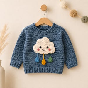 Puede incluir: Un suéter de ganchillo azul de manga larga con una aplicación central de nube color crema que presenta una cara sonriente, mejillas rosadas y tres acentos colgantes en forma de gota en azul, amarillo y verde.