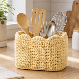May include: A rectangular, pale yellow crochet basket with a scalloped top edge. The basket contains a wooden spoon, a wooden spatula, a metal fork, a metal spoon, and a metal wire whisk, all standing upright within the textured yarn container.