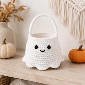 May include: A white crocheted basket shaped like a ghost with a single handle. The basket features two small black circular eyes and a small black curved mouth stitched onto the front. The bottom edge has a scalloped design.