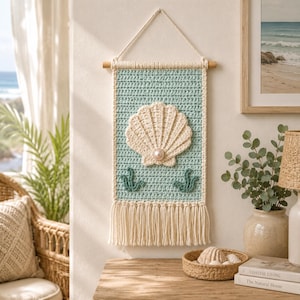May include: A rectangular crochet wall hanging featuring a light blue background with a large cream-colored seashell at the center, accented by a single pearl bead. The piece includes two small green seaweed motifs and a cream fringe at the bottom.