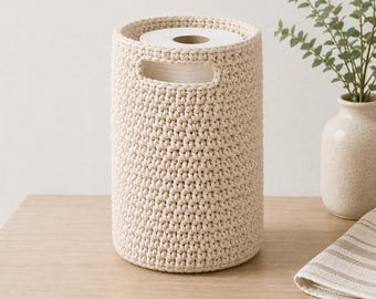 May include: A cylindrical, cream-colored crocheted basket with a textured, woven stitch pattern. The basket features two integrated oval-shaped handles near the top rim and contains a single roll of white toilet paper inside the open center.