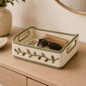 Puede incluir: Una bandeja de almacenamiento rectangular de ganchillo en color crema con bordes y asas en verde salvia. El exterior presenta un patrón de enredadera bordado en verde salvia. Dentro hay unas gafas de sol marrones, unas llaves y una libreta.