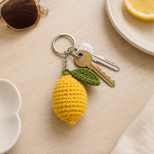 May include: A yellow crocheted lemon keychain with a small green leaf attached to a silver metal split ring. Two metal keys are attached to the ring. The lemon has a textured, yarn-based surface and is shaped like a small, rounded citrus fruit.