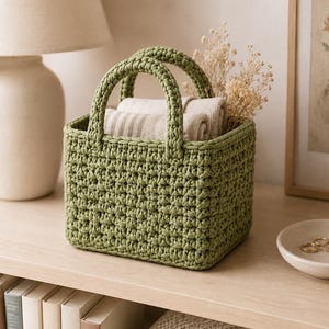 May include: A rectangular sage green crochet basket featuring two sturdy handles and a textured stitch pattern. The basket contains folded beige fabric and dried floral stems, showcasing a structured design with a flat base and vertical sides.