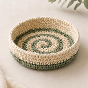 May include: A round, shallow basket made of crocheted cord featuring a spiral pattern in alternating cream and sage green colors. The basket has a thick, textured wall and a flat base, with the green and cream yarn woven in a continuous circular design.