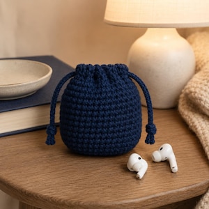 May include: A small, dark navy blue crochet drawstring pouch with a textured, knotted stitch pattern. The pouch features two matching dark navy blue braided cords that cinch the top closed, with knotted ends hanging down on both sides of the bag.