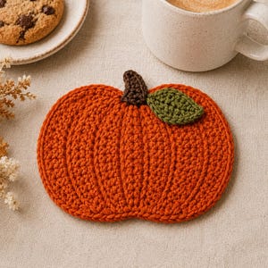 May include: A pumpkin-shaped crochet coaster in vibrant orange yarn with a dark brown stem and a single green leaf. The coaster features a textured ribbed design and measures 6 inches by 5 inches as indicated by the printed text below it.