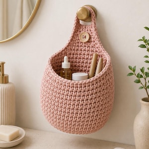 May include: A dusty pink crocheted hanging basket made of thick cord, featuring a single wooden button closure at the top. The basket is suspended from a round brass wall hook and contains a small glass dropper bottle, a white jar, and pens.