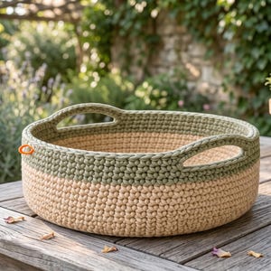 May include: A round, two-toned crochet basket featuring a beige base and a sage green upper section with two integrated handles. The basket has a thick, textured stitch pattern and a small orange loop attached to the side rim for hanging or carrying.