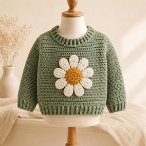 Op de afbeelding: Een saliegroene trui met lange mouwen en een grote, gestructureerde, gehaakte madeliefjesapplicatie op de voorkant. De bloem heeft witte bloemblaadjes en een goudgeel hart. De trui is voorzien van een geribbelde ronde hals, boorden en zoom.