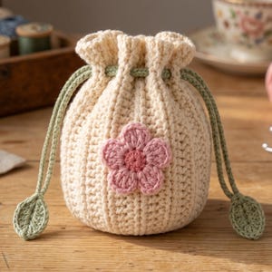 May include: A cream-colored crochet drawstring pouch featuring a ribbed texture and a central pink flower appliqué. The bag is cinched with sage green cords that terminate in small, crocheted leaf-shaped accents at each end of the drawstring.