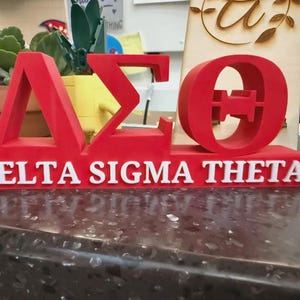 May include: A red three-dimensional sign featuring the Greek letters Delta, Sigma, and Theta in a bold font. Below the Greek letters, the words DELTA SIGMA THETA are displayed in white, raised, capital letters on a red rectangular base.
