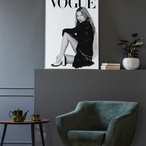 Puede incluir: Portada de la revista Vogue en blanco y negro con una modelo sentada con un vestido negro y tacones. La revista se muestra en una pared gris sobre un sill&oacute;n verde oscuro y una peque&ntilde;a mesa con una tetera y una taza.