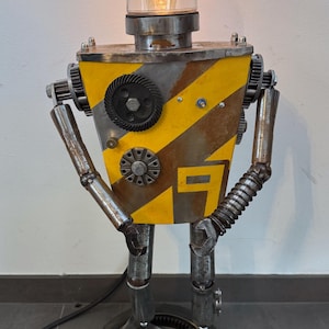 Op de afbeelding: Een handgemaakte robotlamp gemaakt van hergebruikte metalen onderdelen. De behuizing is geel met diagonale zwarte strepen. Het heeft een heldere glazen lampenkap en een warm licht. De lamp is ongeveer 40 cm hoog.