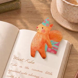 May include: A die-cut paper bookmark shaped like an orange squirrel wearing a teal party hat with pink polka dots. The squirrel is holding a pink gift box with teal stripes. The bookmark is placed on an open book with the text Meet Kiko printed on it.
