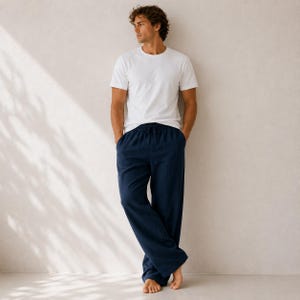 May include: A plain white short-sleeved crew neck t-shirt paired with loose-fitting navy blue lounge pants featuring an elastic waistband with a drawstring tie. The clothing set is worn by a person standing against a neutral light-colored wall.