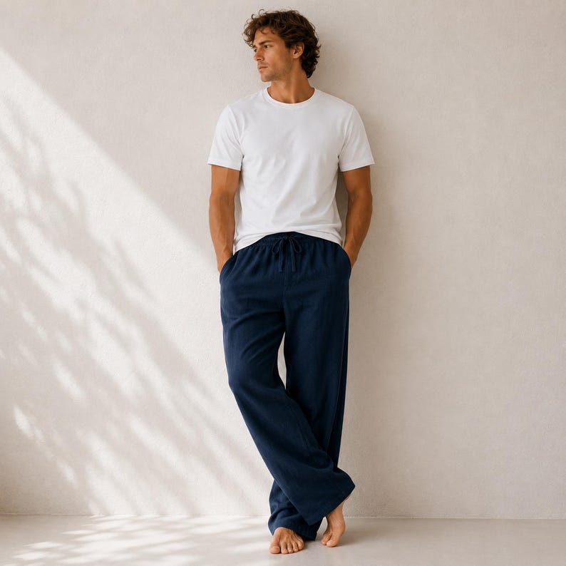 May include: A plain white short-sleeved crew neck t-shirt paired with loose-fitting navy blue lounge pants featuring an elastic waistband with a drawstring tie. The clothing set is worn by a person standing against a neutral light-colored wall.