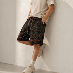 May include: Knee-length shorts featuring a dark-toned geometric pattern in shades of red, green, and blue. The shorts include a drawstring waistband, side pockets, and a solid black hemline detail, paired with a plain beige t-shirt and sneakers.