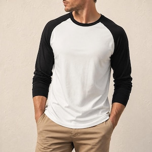 Könnte beinhalten: Ein langärmeliges Baseball-Shirt mit weißem Rumpf und kontrastierenden schwarzen Raglanärmeln sowie einem Rundhalsausschnitt. Das Shirt wird mit einer beigefarbenen Hose kombiniert und zeichnet sich durch einen klassischen Schnitt aus.