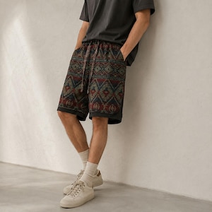 May include: Knee-length shorts featuring a dark-toned geometric pattern in shades of red, blue, and green. The shorts have an elastic waistband with a matching drawstring, paired with a plain dark gray t-shirt, white crew socks, and white sneakers.