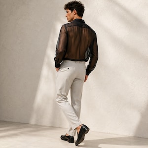 May include: A long-sleeved black sheer button-down shirt with a collar and cuffs, paired with light gray trousers featuring a single visible button at the back waist, and black leather loafers worn on the feet, captured from a rear-facing angle.