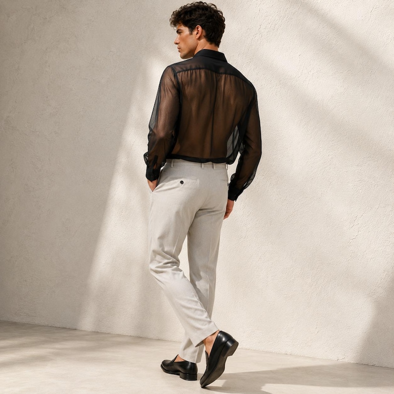 May include: A long-sleeved black sheer button-down shirt with a collar and cuffs, paired with light gray trousers featuring a single visible button at the back waist, and black leather loafers worn on the feet, captured from a rear-facing angle.