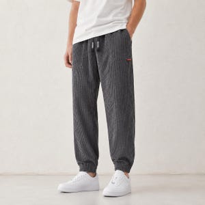 Puede incluir: Pantalones de pana gris carbón oscuro con textura de rayas verticales, cintura elástica con cordón, dos bolsillos laterales y puños en los tobillos. Se observa una pequeña etiqueta rectangular roja en la zona del bolsillo derecho.