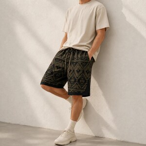 May include: A plain beige short-sleeved t-shirt paired with black shorts featuring a dark olive green geometric pattern. The shorts include a drawstring waistband and a solid black hemline. The outfit is completed with white athletic sneakers.