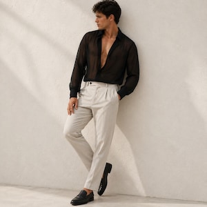 May include: A long-sleeved black shirt made of a sheer, translucent fabric with a deep V-neckline, paired with light beige trousers featuring a pleated front, a button closure, and a straight-leg cut, worn with black leather slip-on loafers.