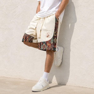 May include: Off-white shorts featuring a drawstring waist with tassel ends and a colorful geometric pattern along the side panels and hem. A small square patch with a graphic design is visible on the front. The shorts are paired with a white t-shirt.