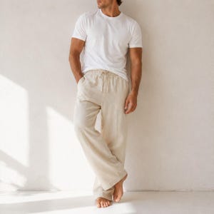 May include: A plain white short-sleeved crew neck t-shirt paired with loose-fitting beige trousers. The trousers feature a drawstring waistband with matching beige ties and a wide-leg cut that falls straight to the ankles, creating a relaxed silhouette.