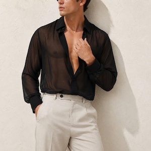 May include: A black long-sleeved shirt made of a sheer, translucent fabric featuring a classic collar, a button-down front, and buttoned cuffs. The shirt is worn unbuttoned at the top and is paired with light beige trousers that have a visible button closure.